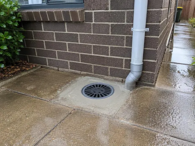 Outdoor drain grate beside brick wall and downpipe with wet paving in Matraville